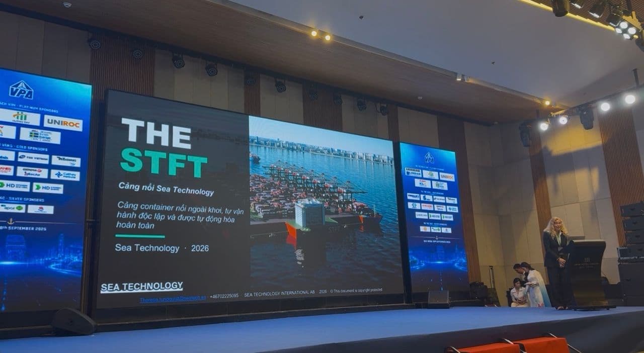 Speaker at a podium on stage with large screens showing slides for THE STFT floating container port by Sea Technology