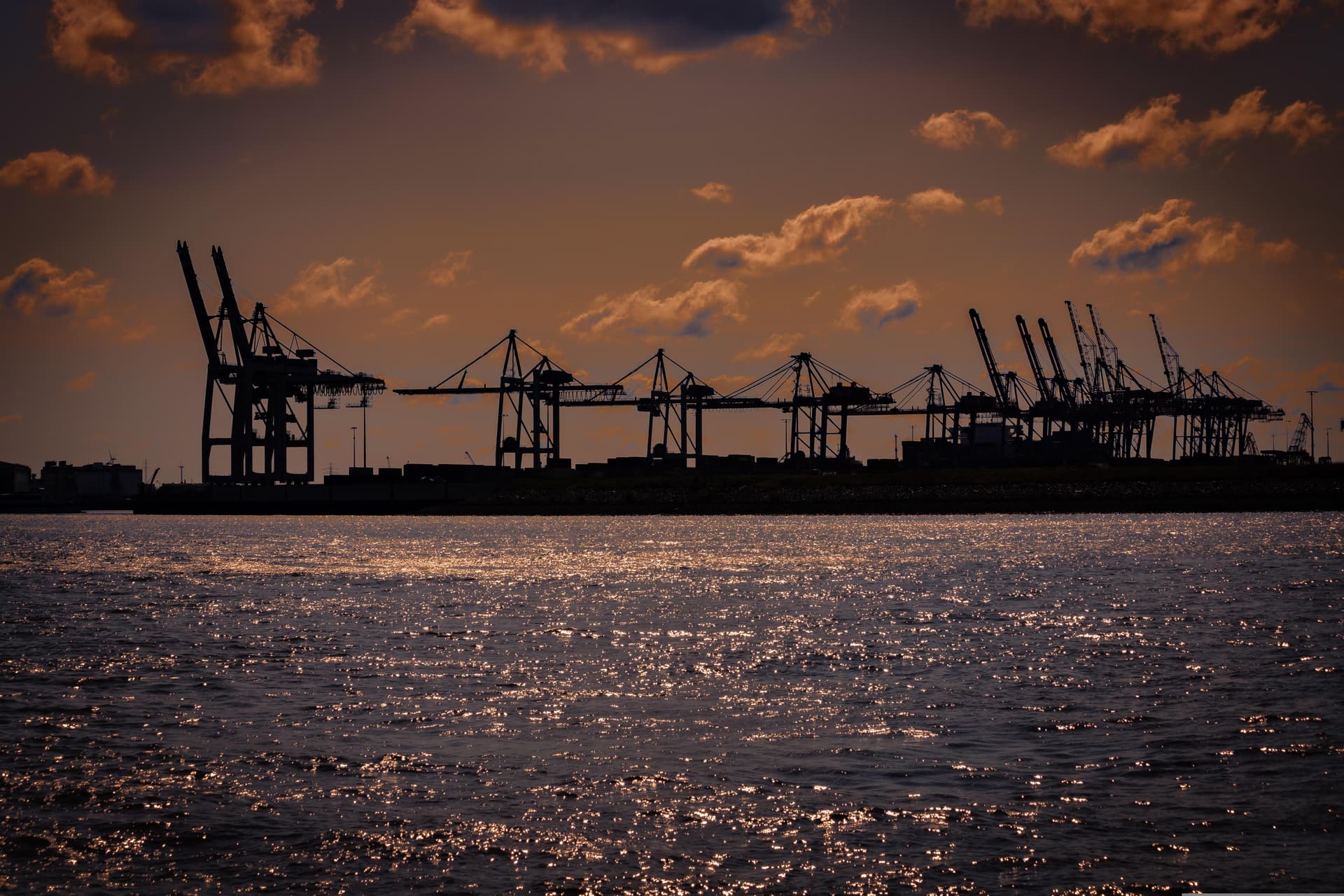 Silhouetted container port cranes at sunset across shimmering water